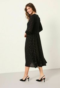Schwarzes Kleid mit goldenem Punktemuster, langen transparenten Ärmeln, fließendem Rock und tailliertem Oberteil. Kombiniert mit schwarzen Slingback-Absätzen.