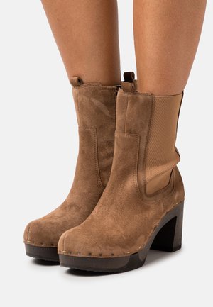 Bottines en daim marron avec un large talon en bois, dotées de panneaux latéraux élastiques et de coutures décoratives le long de la semelle.