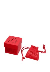 Boîte carrée rouge avec de petits trous sur le dessus, portant le logo "DIESEL" en relief. Accompagnée d'un sac à cordon rouge assorti.