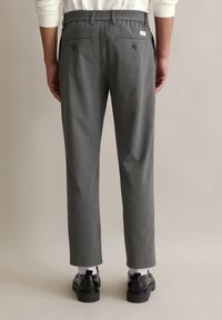 Grijze geruite broek met een elastische tailleband, twee achterzakken met knopen, taps toelopende pijpen, gedragen met zwarte schoenen en witte sokken.