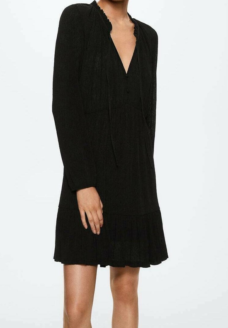 Femme portant une robe noire à manches longues avec un col en V et un ourlet plissé, montrée du cou jusqu'à mi-cuisse sur un fond uni.