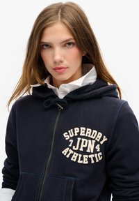Marineblå lynlås hættejakke med snoretræk, featuring en front kangaroo lomme og hvid tekst, der siger "SUPERDAY JPN ATHLETIC." Blød tekstur.