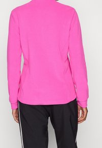 Přiléhavý dlouhý rukáv top ve sytě růžové barvě, vyrobený z měkké látky s kulatým výstřihem a plochými švy. Kombinován s černými kalhotami s bílými pruhy.