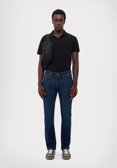 Black polo shirt, blue denim jeans, green sneakers. Model wears a black crossbody bag. Fabrics are smooth; jeans have a classic fit.