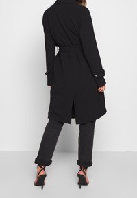 Musta trench-takki, jossa on vyötärölle sidottu vyö, pitkät hihat ja takakäytävä. Yhdistetty tummiin lyhyisiin farkkuihin ja nilkkaremmikenkiin.