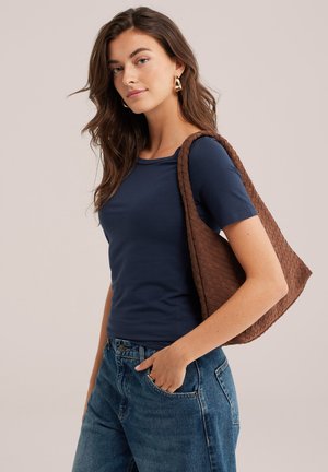 Femme aux longs cheveux bruns portant un t-shirt bleu marine à manches courtes et un jean, avec un sac à bandoulière tressé marron et des boucles d’oreilles créoles dorées.