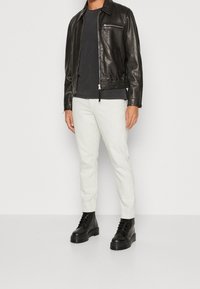 Giacca di pelle nera con tasca con zip, abbinata a una maglietta grigia e pantaloni slim-fit di colore chiaro. Stivaletti neri con suola spessa.