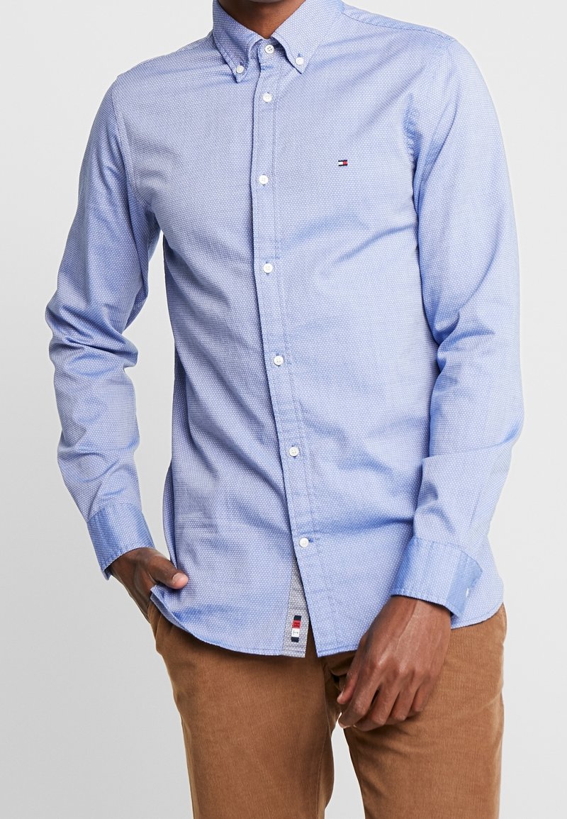 Homme portant une chemise boutonnée à manches longues bleu clair à motifs et un pantalon en velours côtelé marron, la main posée dans la poche.
