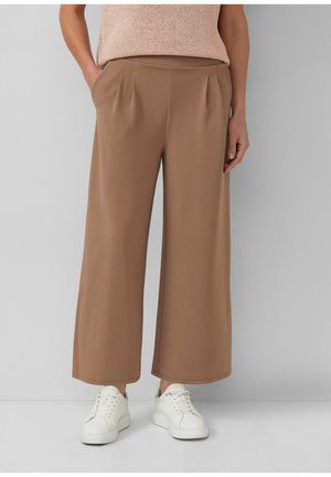 Personne portant un pantalon marron taille haute à jambes larges avec poches latérales, associé à des baskets blanches et un haut beige sans manches.