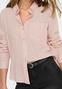 Camisa de botones rosa claro con un bolsillo, mangas largas remangadas, hecha de tejido suave, combinada con jeans negros y un cinturón negro.