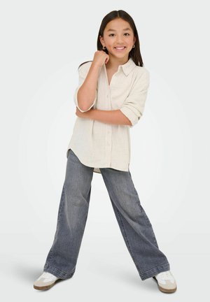 Une fille souriante se tient les jambes écartées, portant une chemise beige à boutons, un jean gris à jambes larges, des baskets blanches et des boucles d'oreilles en or.