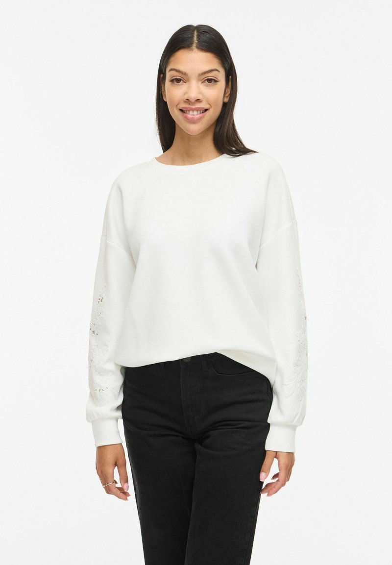 Sieviete ar garām tumšām matiem valkā baltu džemperi ar ziedu izšuvumiem uz piedurknēm un melnas augsta vidukļa bikses, stāvot pret baltu fonu.