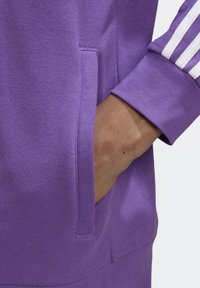 Lila sweatshirt med en sidficka och kontrasterande vita ränder på ärmslutet. Tyget verkar mjukt med en normal passform.