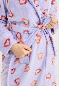 Lavendelkleurige fleecebadjas met oranje hartjespatroon. Voorzien van een ceintuur, zakken en lange mouwen. Zachte textuur met een ontspannen ontwerp.
