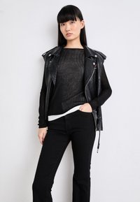 Svart skinnvest med sølvfarget hardware over en gjennomsiktig svart topp, lagdelt med hvit kant. Modellen har på seg svarte vide jeans, med hendene i lommene.