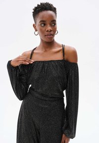 Zwarte off-shoulder top met lange mouwen, voorzien van een aansluitende taille en een sprankelende textuur. Dunne, versierde bandjes voegen detail toe aan het ontwerp.