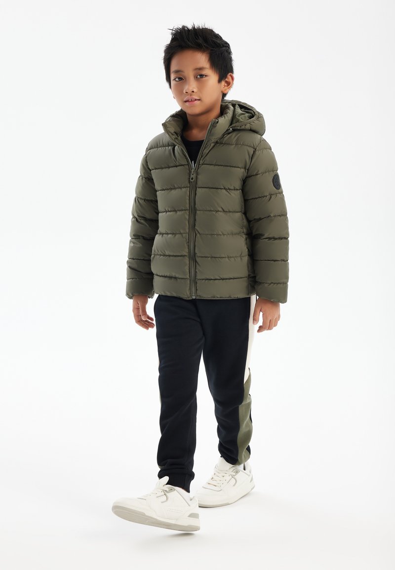 Veste rembourrée verte avec capuche, fermeture éclair et design matelassé. Associée à un pantalon noir avec une bande d'accent verte et des baskets blanches.