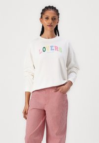 Fehér pulóver színes "LOVERS" felirattal és alatta "Self Care Club", piros-fehér csíkos nadrággal párosítva. Kényelmes fazon, puha anyag.
