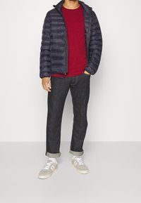 Marinblå puffjacka, röd t-shirt med rund hals, mörka jeans och grå sneakers med vita detaljer. Har horisontellt kviltat mönster och uppvikta ärmar.