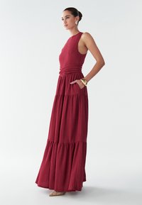 Kvinde i en ærmeløs, lagdelt, tekstureret rød maxikjole med hånden i lommen, iført guldarmbånd, øreringe og dekorerede spidse sko.