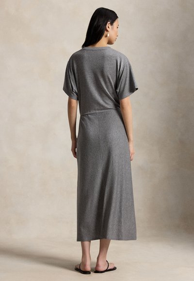 Mujer de pie de espaldas, vistiendo una blusa gris suelta de manga corta y una falda larga a juego, con sandalias negras sobre un fondo neutro.