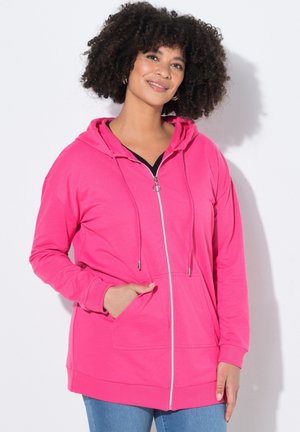 Frau mit lockigem Haar, die eine leuchtend pinke Kapuzenjacke mit Reißverschluss trägt, mit der linken Hand in der vorderen Tasche, lächelnd vor einem schlichten weißen Hintergrund.