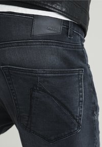 Vaqueros de denim oscuro con ajuste slim, costuras en contraste y una distintiva etiqueta de cuero con el nombre de la marca "CHASIN." en la cinturilla.