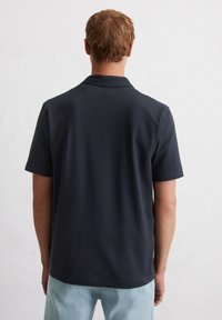 Man met kort lichtbruin haar, gekleed in een donkerblauw shirt met korte mouwen en lichtblauwe jeans, staand met zijn gezicht naar een witte muur.