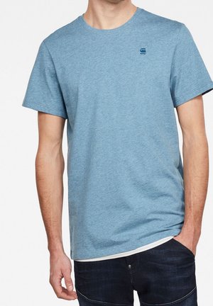 Homme portant un t-shirt bleu clair à manches courtes avec un petit logo sombre sur la poitrine et un jean bleu foncé, main dans la poche.
