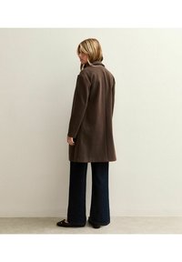 Abrigo marrón de mezcla de lana con cuello de muesca, mangas largas y un corte relajado, llevado sobre pantalones acampanados de mezclilla oscura y zapatos de charol negros.