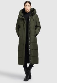 Cappotto imbottito verde oliva con cappuccio, lunghezza lunga, design trapuntato, dettagli neri e tasche laterali, abbinato a stivali neri fino al ginocchio.