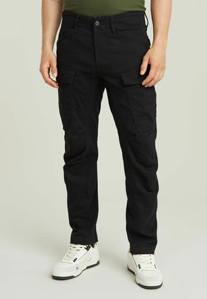 Homme portant un pantalon cargo noir avec des poches latérales et des baskets blanches, debout sur un fond clair uni.