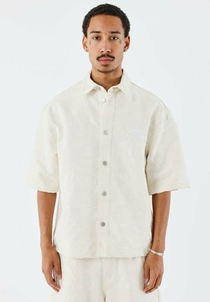 Chemise crème à manches courtes avec un motif floral texturé, des boutons argentés et un détail de logo bleu sur la poitrine gauche.