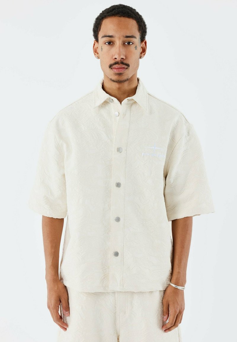 Crème short-sleeve shirt met een текстурное bloemmotief, zilveren knopen en een logo in het blauw op de linkerborst.