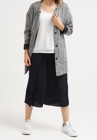 Cárdigan gris y blanco con estampado, con ribete negro, sobre una blusa blanca, combinado con culottes marinos y zapatillas blancas. Manga larga, corte relajado.