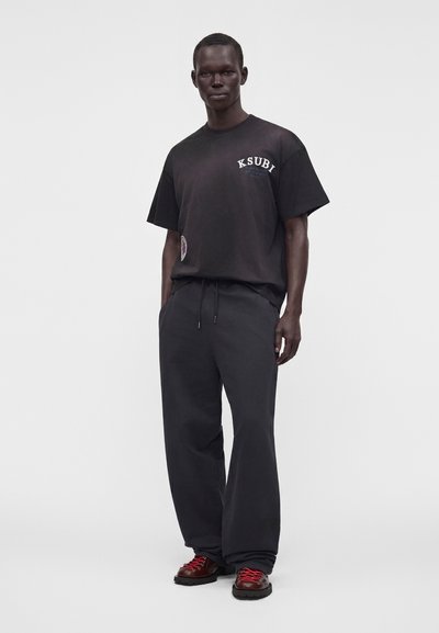 Άνδρας με μαύρο t-shirt Ksubi, φαρδιά μαύρη παντελόνια και σκούρα καφέ παπούτσια με κόκκινα κορδόνια, στέκεται μπροστά σε απλό λευκό φόντο.