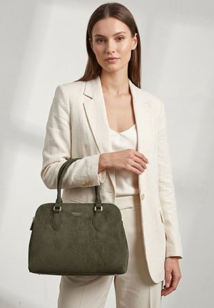 Femme en tailleur crème tenant un sac à main structuré vert foncé avec des doubles poignées, debout devant un fond clair uni.