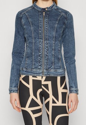 Vrouw draagt een getailleerd blauwe spijkerjack met ritssluiting aan de voorkant en zwarte legging met beige abstracte geometrische patronen.