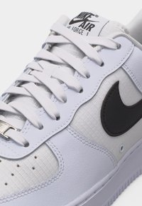 Gros plan sur une basket Nike Air Force 1 blanche avec le logo swoosh noir, des lacets blancs et une matière supérieure texturée.