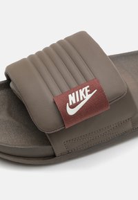 Barna csúszó szandál párnázott pánttal, függőleges bordákkal, burgundi Nike logóval és texturált lábbelivel a tapadás érdekében.