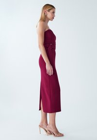 Bordo brez naramnic obleka z oprijetim zgornjim delom, stranskim razporkom in pasom iz tkanine. Gladek teksturni material dolžine do sredine meč. Nošena z bež sandali na peto.