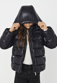 Vaikas, vilkintis juodą pūkinį švarką su kapucėle, kuri dalinai dengia veidą, abu rankas laiko už kapučio kraštų, vilkintis baltą marškinėlį ir juodas kelnes.