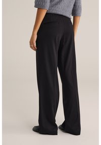 Fekete, bő szárú nadrág sima textúrával, két visszafogott hátsó zsebbel, szürke kötött felsővel párosítva.