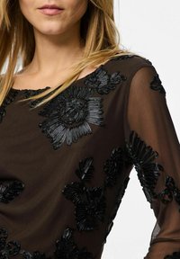 Mustast läbipaistvast kangast pluus, millel keerukas lilleline tikand läikivate detailidega, ümara kaelusega ja pikad varrukad. Pehme, kerge kangas.