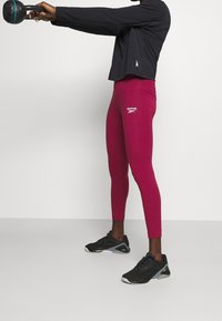 Black long-sleeve top, fitted maroon leggings with Reebok logo, and black training shoes with gray accents; weights held out front.