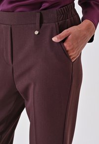 Pantaloni bordeaux con una texture liscia, dotati di una vita elasticizzata e di una tasca frontale, chiusi con un bottone argentato.