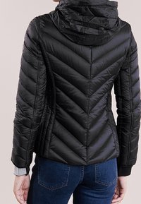 Sort dunjakke med hætte, præget af en chevron-quiltet mønster, glat tekstur og ribbede manchetter. Bæres med jeans.