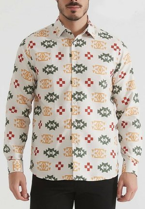 Homme portant une chemise blanche à manches longues avec des motifs géométriques rouges, verts et jaunes, associée à un pantalon noir.