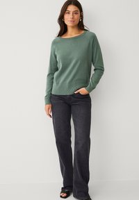 Maglione verde a maglia con scollatura ampia, polsini e orlo a coste, abbinato a jeans svasati scuri e sandali neri con punta aperta.