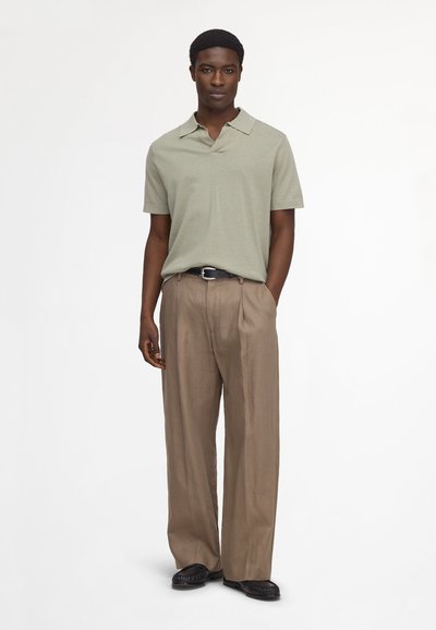 Uomo in piedi con polo verde chiaro a maniche corte, pantaloni beige larghi con cintura nera e scarpe nere su sfondo bianco.